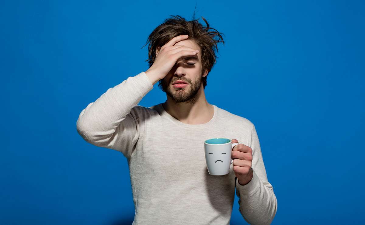 Uomo con espressione stanca che si tiene la testa mentre tiene in mano una tazza con una faccina triste, simbolo del Blue Monday.