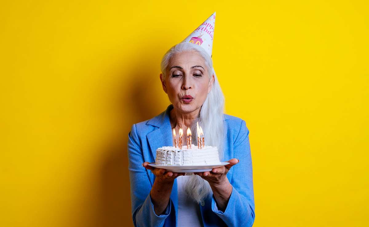 Donna elegante con lunghi capelli grigi e cappellino da festa soffia sulle candeline di una torta di compleanno bianca, su sfondo giallo vivace.