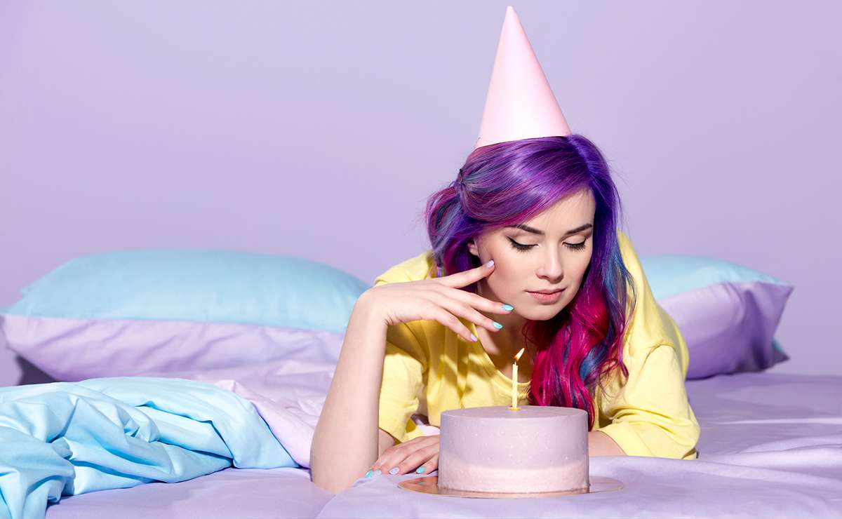 Ragazza con capelli colorati e cappellino da festa osserva una torta di compleanno lilla con una sola candela accesa, in un ambiente pastello.