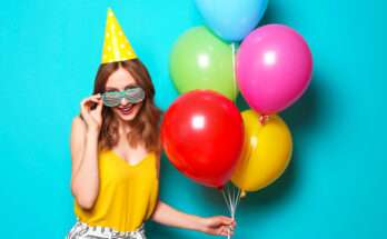 Donna sorridente con cappellino da festa giallo a pois, occhiali divertenti e palloncini colorati su sfondo azzurro, in un’atmosfera festosa di compleanno.